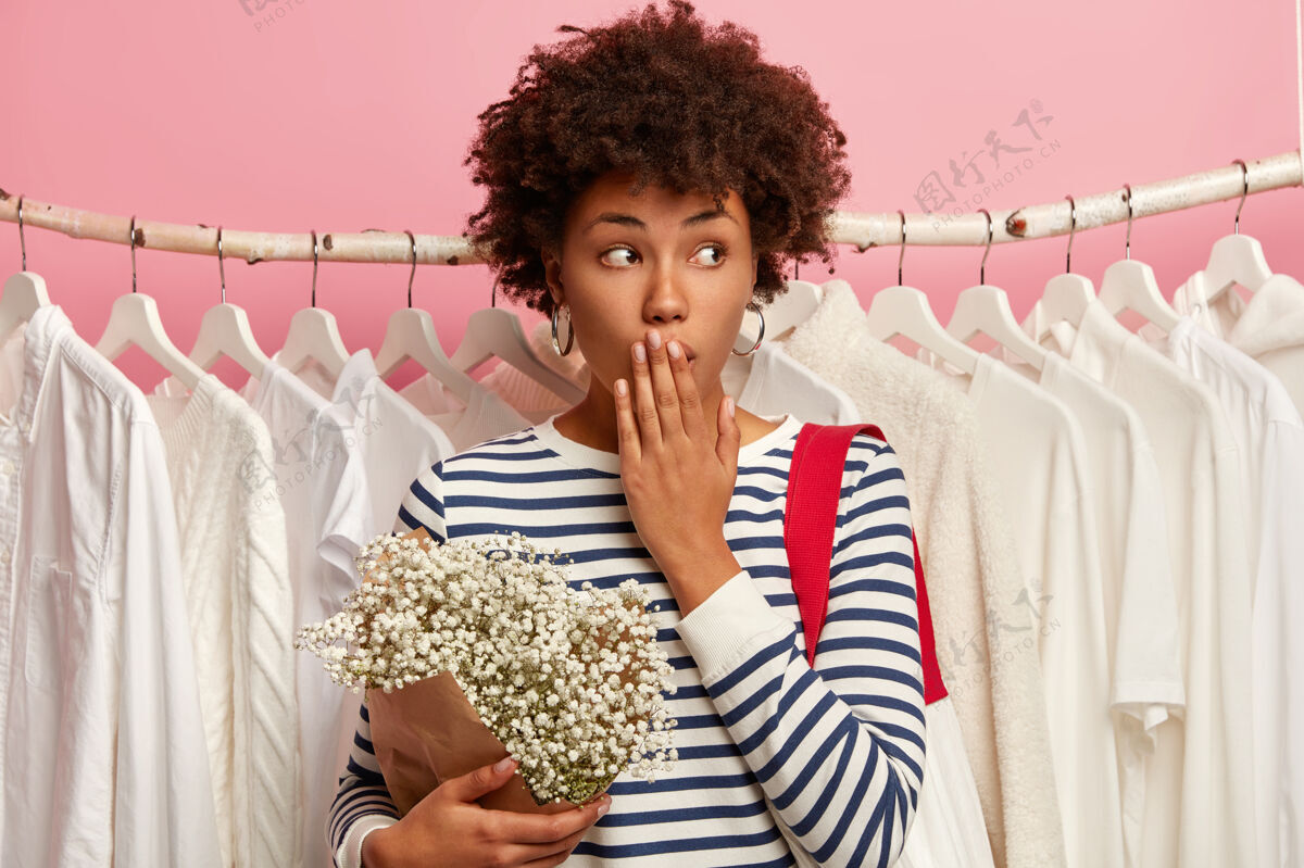 一旁惊讶的非洲裔美国女买家的照片捂着嘴 看向一边 穿着条纹衣服 拿着花束 站在栏杆上一排悬挂着白色的衣服 孤立在粉色上尴尬商店白色