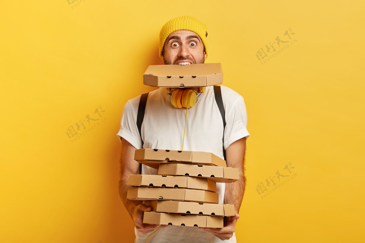 一个超载披萨男的画像拿着许多纸板包裹 一个在嘴里 穿着休闲的白色t恤包装黄色许多