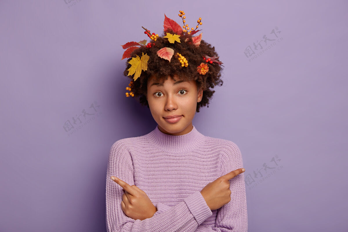 休闲年轻困惑的非洲女人侧身指了指 在两个选项中选择 请求帮助做决定 表示左右 有卷发的秋叶和浆果 孤立在紫色的墙上叶子秋天紫色