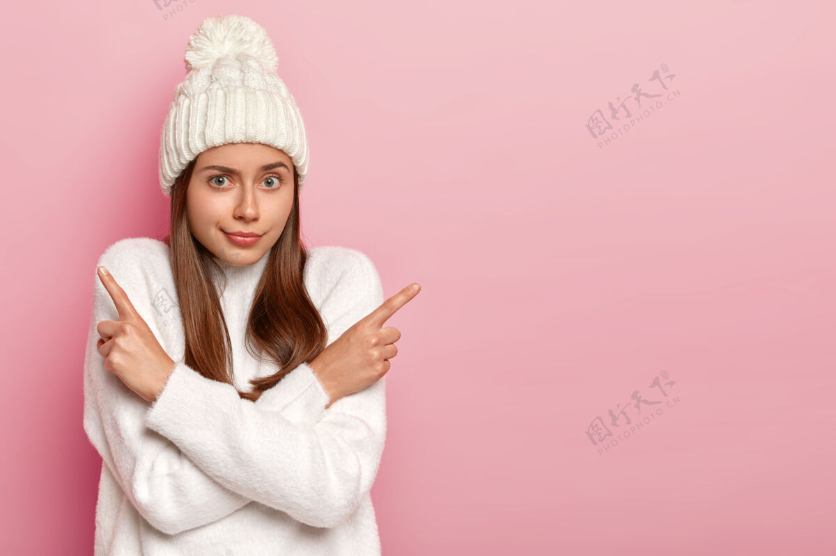指向选择哪一个犹豫不决的自然的欧洲女人侧指 双臂交叉在胸前 建议两个变种 做决定 穿着温暖的白色毛衣和帽子 粉红色背景上的空白帽子女士促销