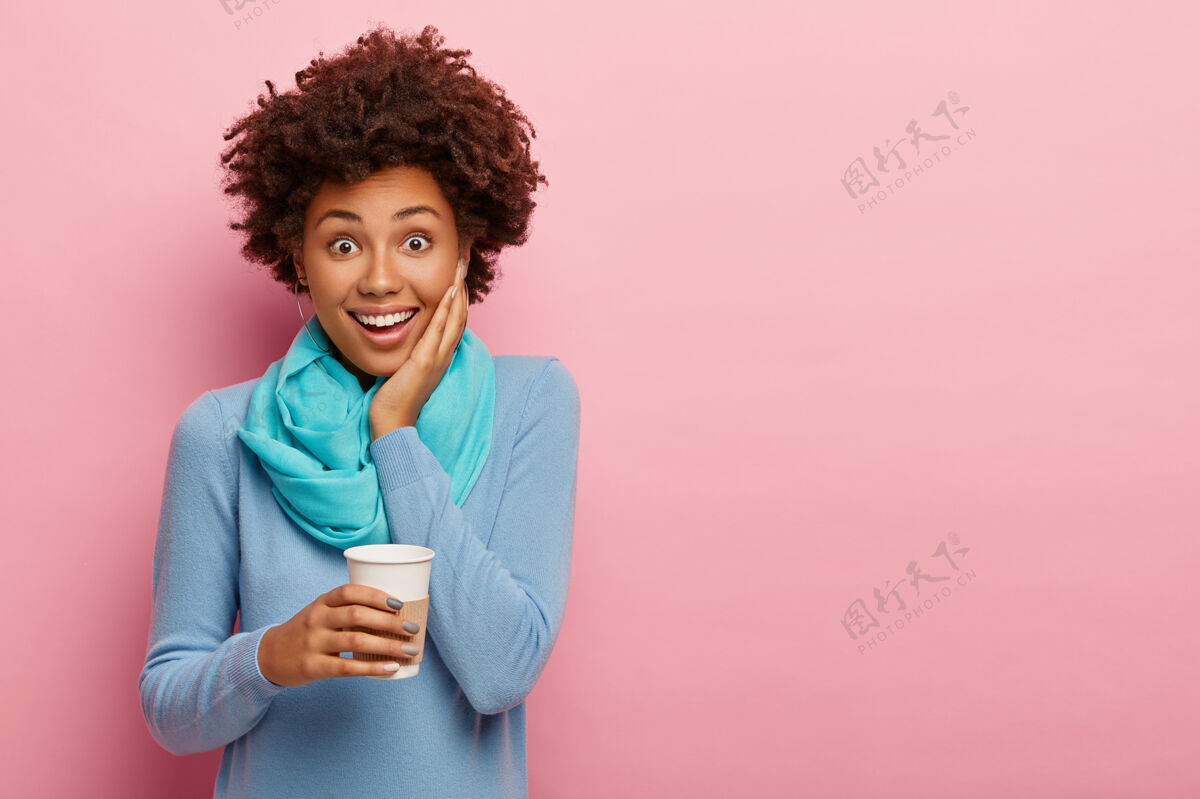 休闲无忧无虑的卷发女人的照片拿着一次性咖啡 喜欢喝芳香饮料 穿着蓝色休闲服 下班后有空闲时间 在玫瑰色的墙上摆姿势 为你的广告复制空间黑发卷发粉色