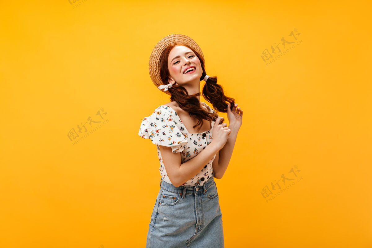 女士扎着两个马尾辫的美女娇媚地笑着戴着帽子 穿着夏装和牛仔裙的女人在橙色的背景下摆姿势牛仔裙印花迷人