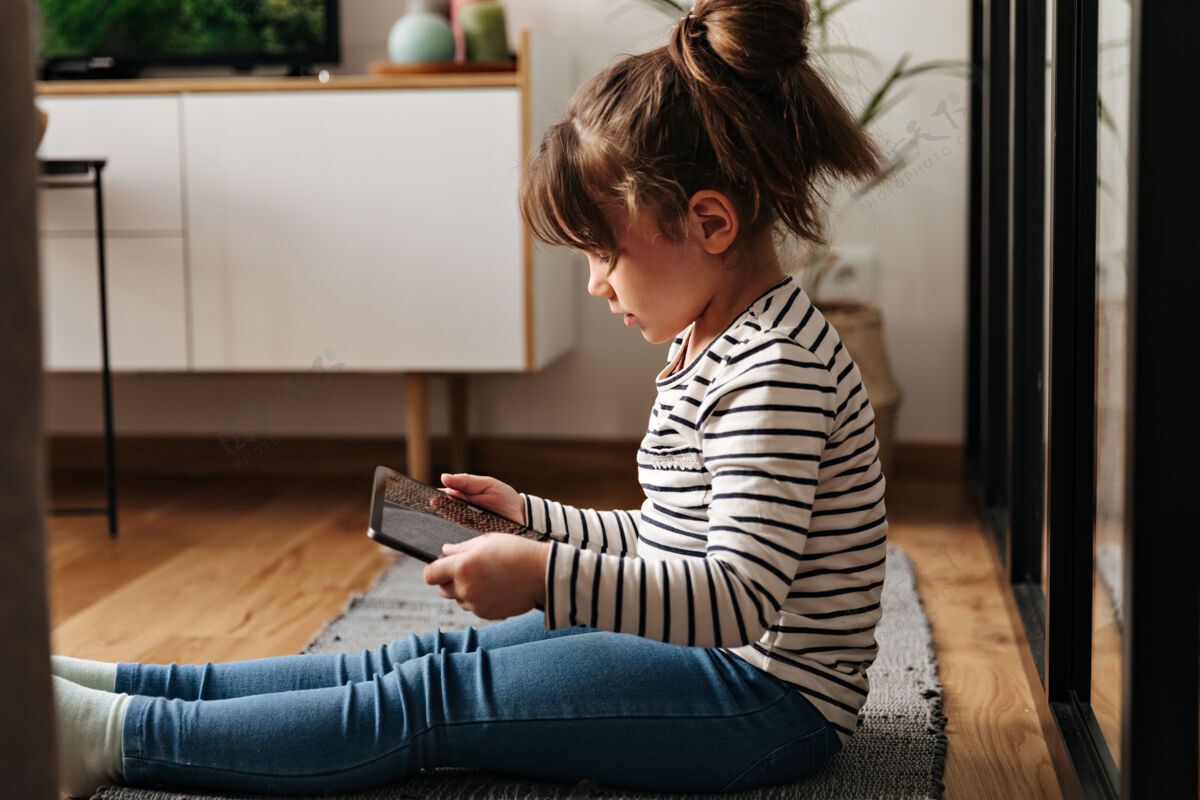 触摸穿着牛仔裤和t恤的小女人坐在地毯上拿着平板电脑的画像朋友科技小