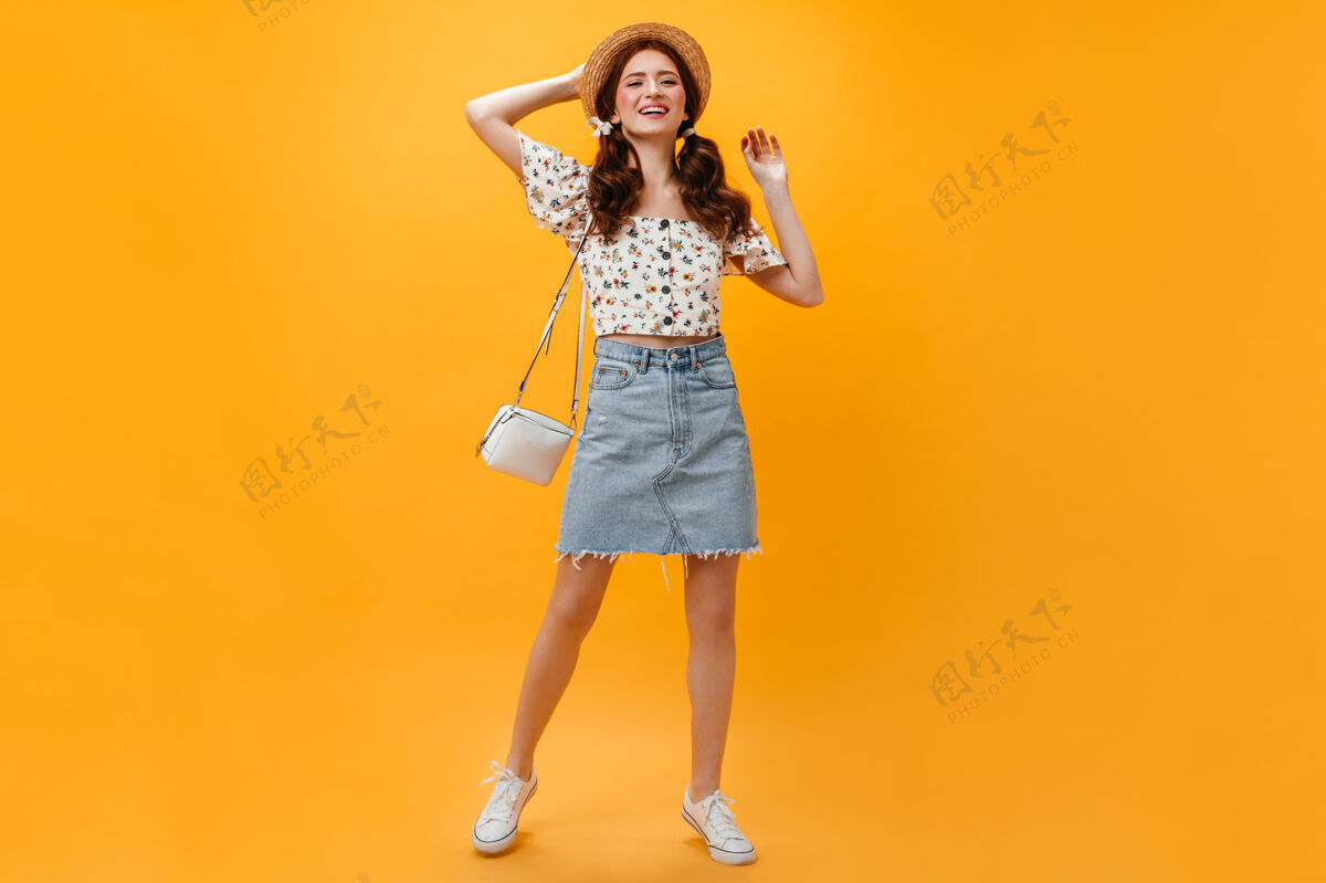 化妆品快乐的女士穿着牛仔裙和不规则剪裁的上衣 在橙色背景上摆出白色包的姿势女人迷人帽子