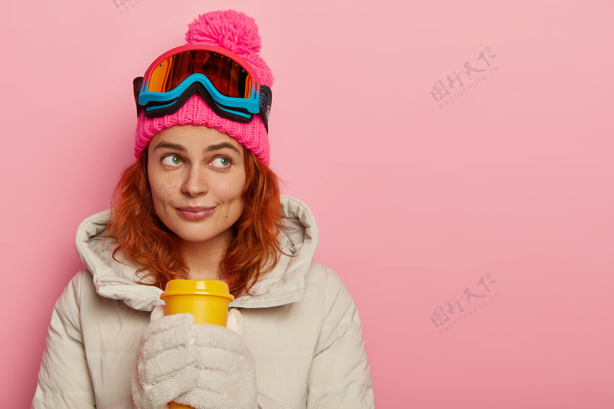 眼镜体贴运动员的女人 穿着暖和的外套 喝外卖咖啡 注意力集中 模特对着粉色的工作室墙女士运动帽子
