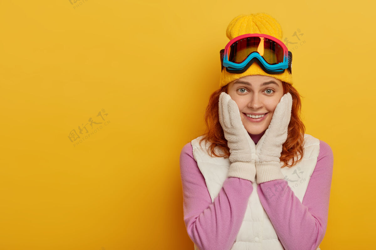 红发可爱快乐的女人双手放在脸颊上 温柔的微笑 微笑着看着镜头 享受着积极的休息和滑雪旅游 穿着积极的衣服 隔离在黄色的墙壁上冬天的时间冬天触摸交叉