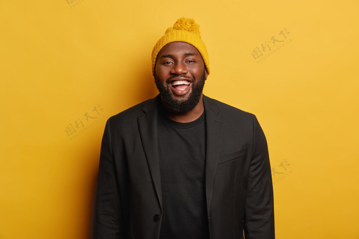 丰满快乐搞笑的男人 穿着黑色西装 戴着黄色冬帽 面带笑容 站在室内鬃毛非洲裔脸