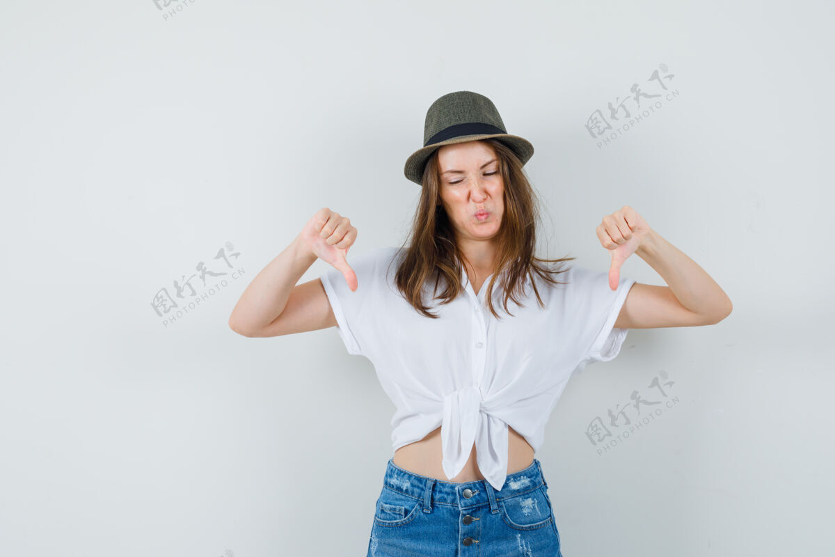 优雅年轻女士在t恤 牛仔裤 帽子上竖起大拇指 看起来很不高兴女人成人向下
