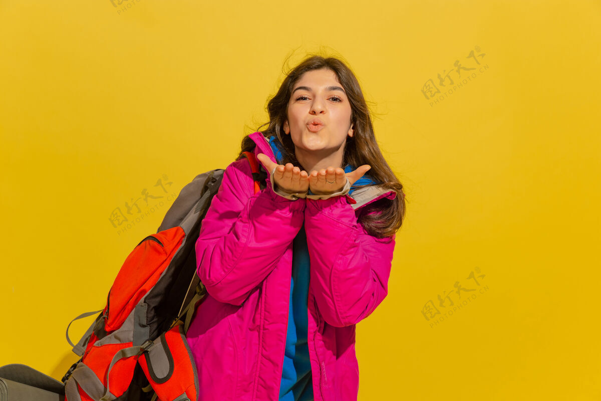 一个微笑着 送上吻一个快乐的年轻高加索旅游女孩的肖像带着包和望远镜隔离在黄色的工作室背景上明亮欢呼喜悦