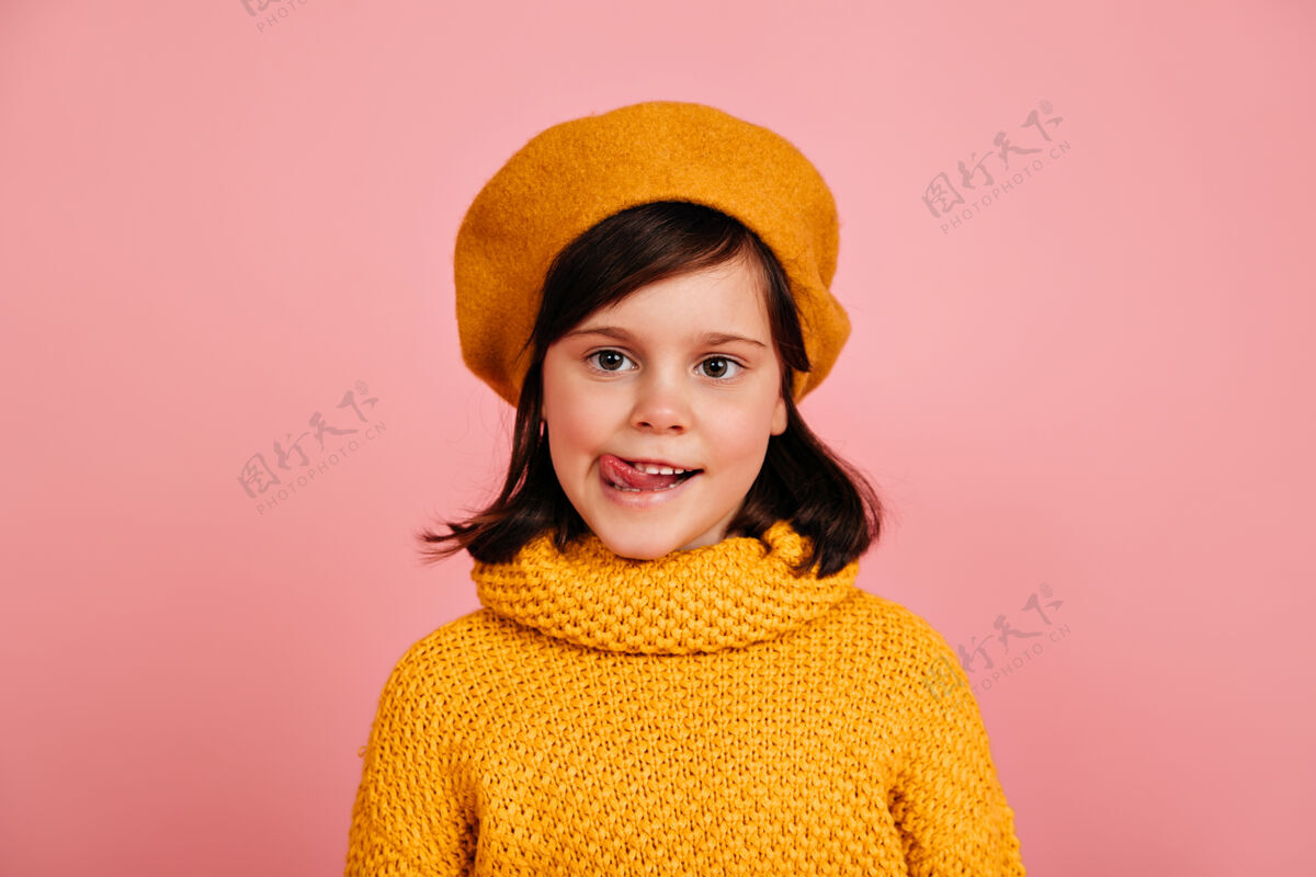休闲穿着针织毛衣的小女孩站在粉红色的墙上孩子摆出舌头的姿势时尚配饰快乐