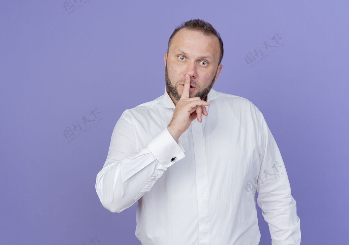 胡子一个留着胡子的男人穿着白衬衫 站在蓝色的墙上做着沉默的手势 手指放在嘴唇上站做男人