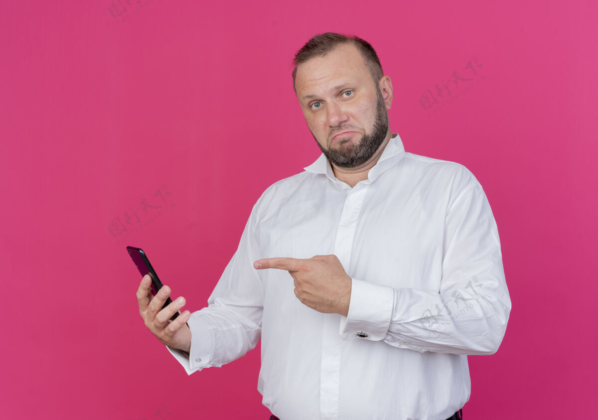 穿一个留着胡子的男人穿着白衬衫 拿着智能手机 用手指着它 站在粉色的墙上 看起来很自信胡子移动自信