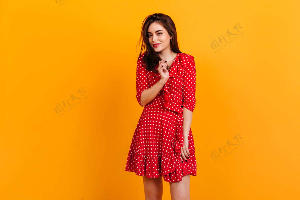 站立穿着红色裙子的时髦女孩的肖像模特在橙色的墙上微笑着黑发颜色头发