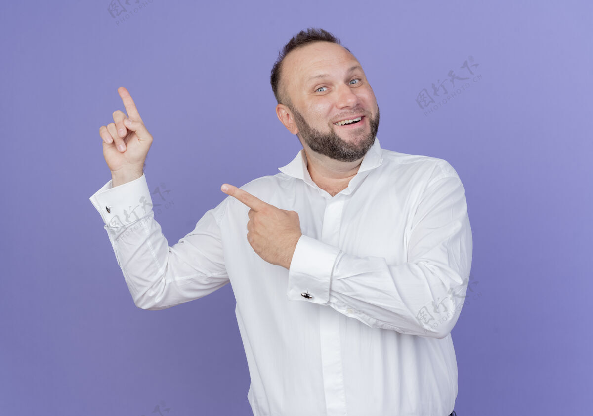 手指满脸胡须的男子身穿白衬衫 面带微笑 用手指着站在蓝色墙上的一边站胡子穿