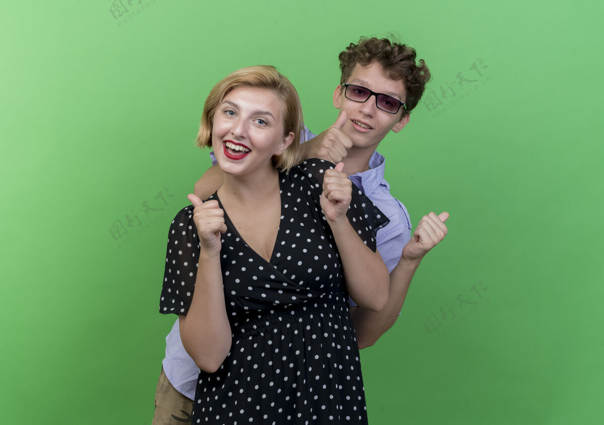年轻年轻漂亮的夫妇站在绿色的墙壁上 微笑着快乐而积极地竖起大拇指女人微笑站
