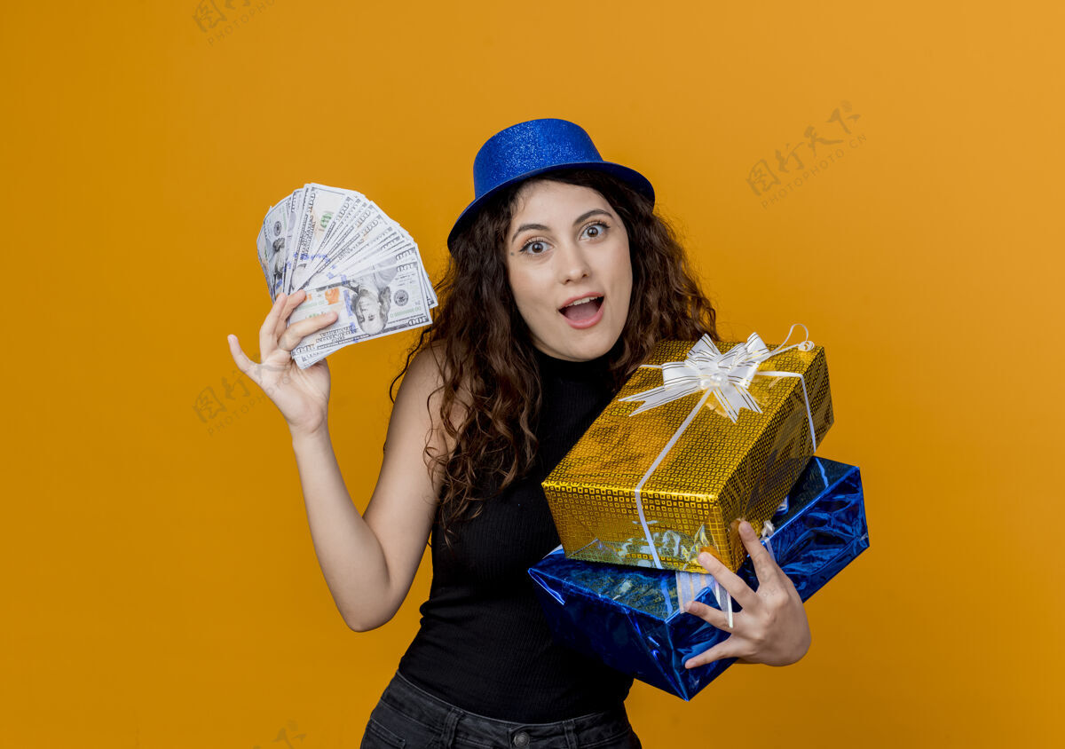 帽子一个年轻漂亮的女人 卷发 戴着派对帽 手里拿着现金和礼物 看着卡内拉站在橘色的墙上 快乐而兴奋卷发抱看