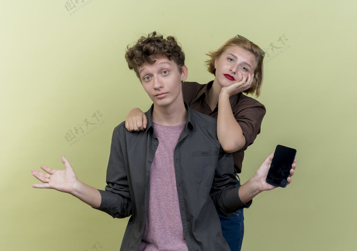 困惑年轻漂亮的夫妇穿着休闲服困惑的男子拿着智能手机耸耸肩和他的女朋友不高兴地站在轻墙不高兴肩耸肩
