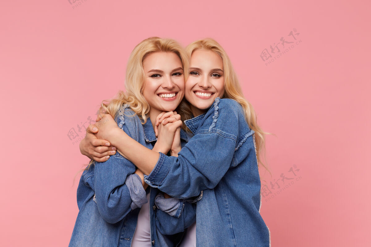 室内摄影棚的照片 年轻的快乐可爱的金发姐妹长波浪发看着相机迷人的微笑 同时拥抱可爱 站在粉红色的背景T恤白色欢呼