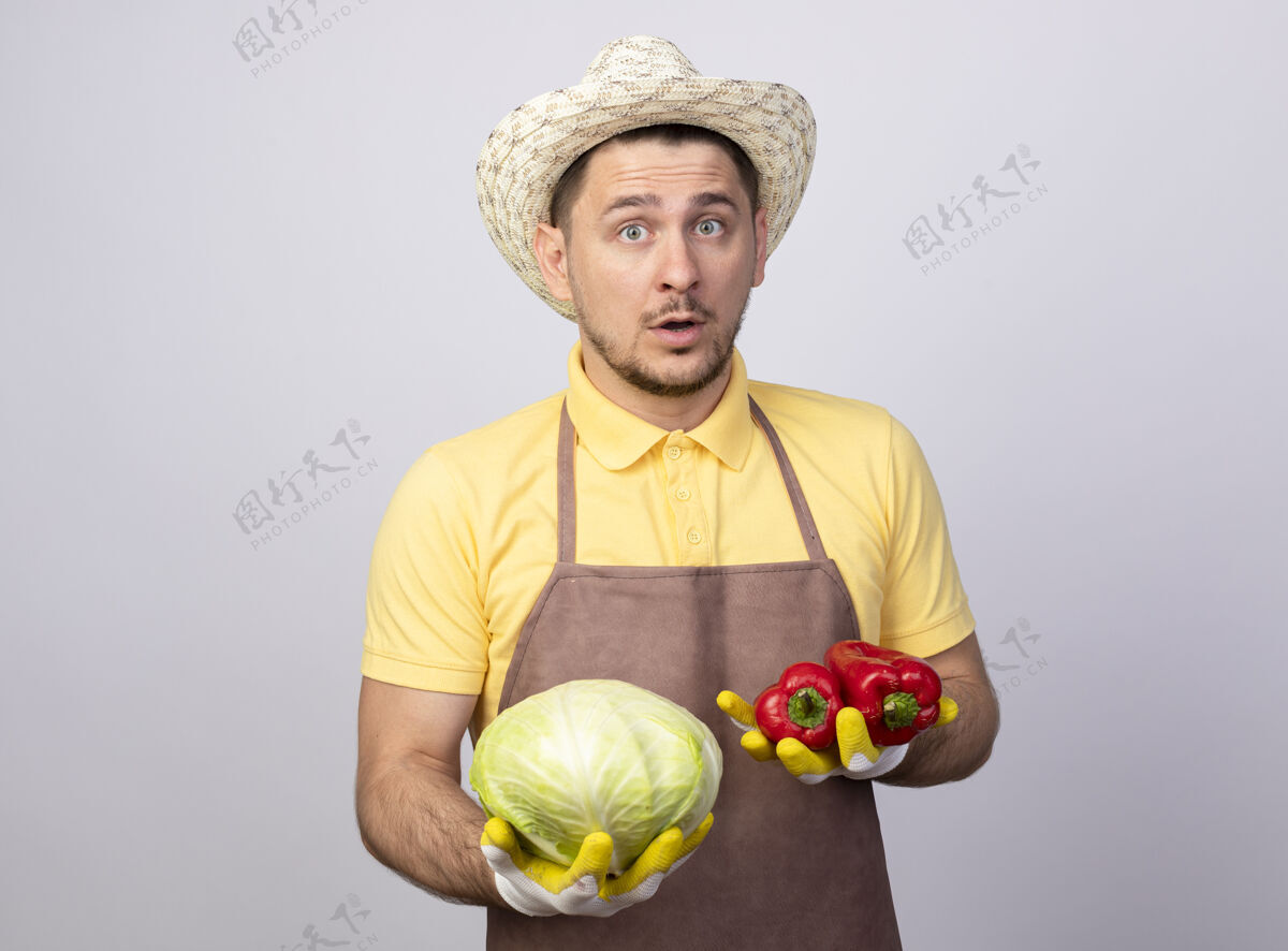 红色年轻的园丁 穿着连体衣 戴着帽子 戴着工作手套 手里拿着红甜椒和卷心菜穿帽子工作