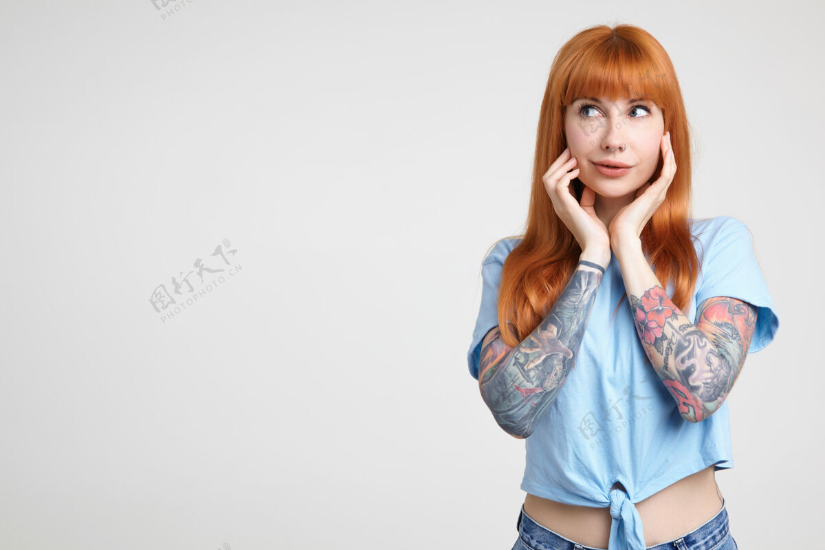 女人年轻漂亮的纹身女性的摄影棚照片 长着一头狐狸精的头发 双手举在脸颊上 一边惊奇地看着一边 隔离在白色背景下女性手红发