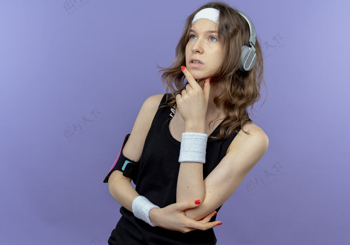 头带身穿黑色运动服 头戴头巾 手戴智能手机臂章的年轻健身女孩站在蓝色的墙上 脸上带着沉思的表情望向一边面部健康移动