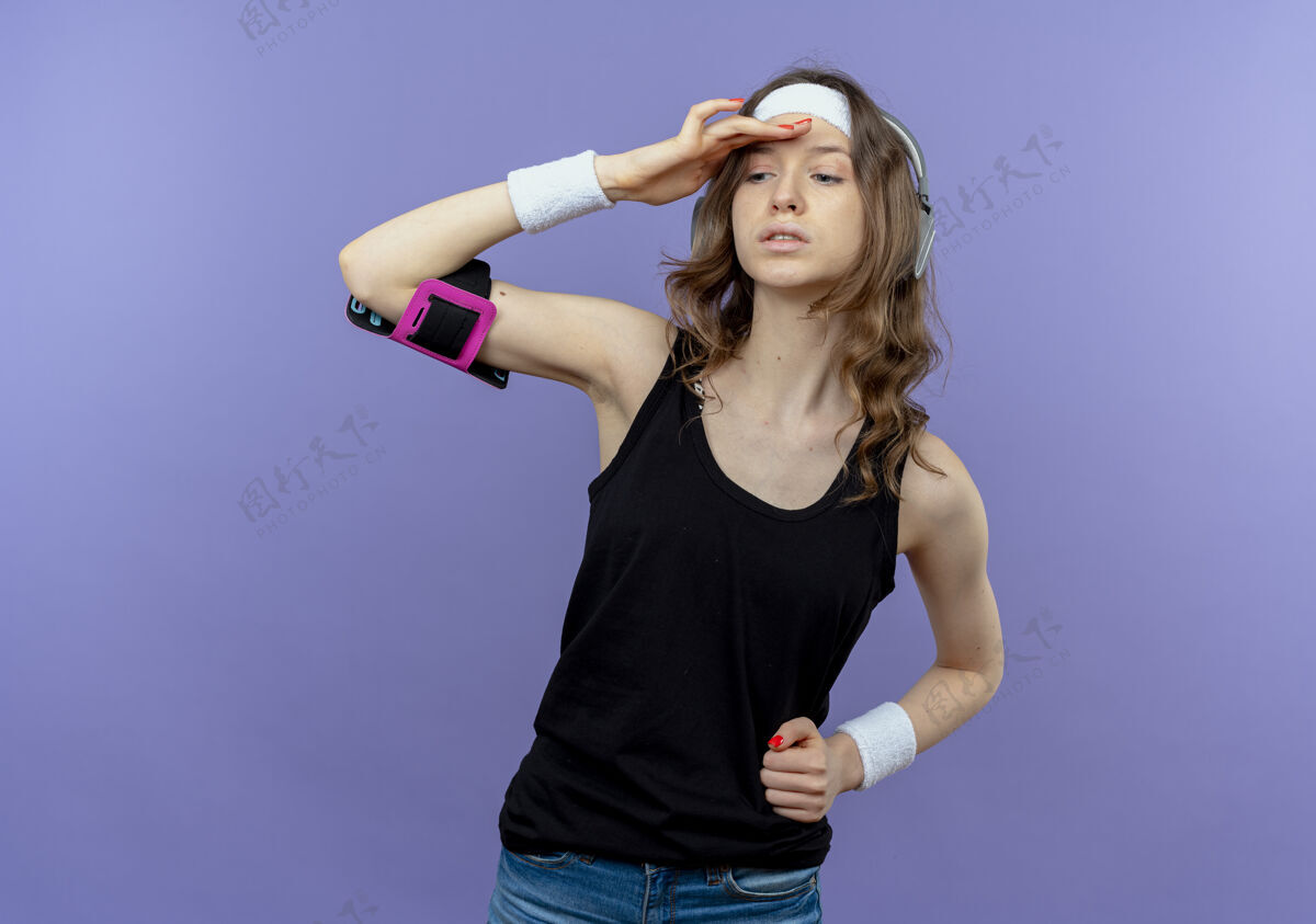 手身穿黑色运动服 头戴头巾 手戴智能手机臂章的年轻健身女孩远远望去 手举着头站在蓝色的墙上头健身远