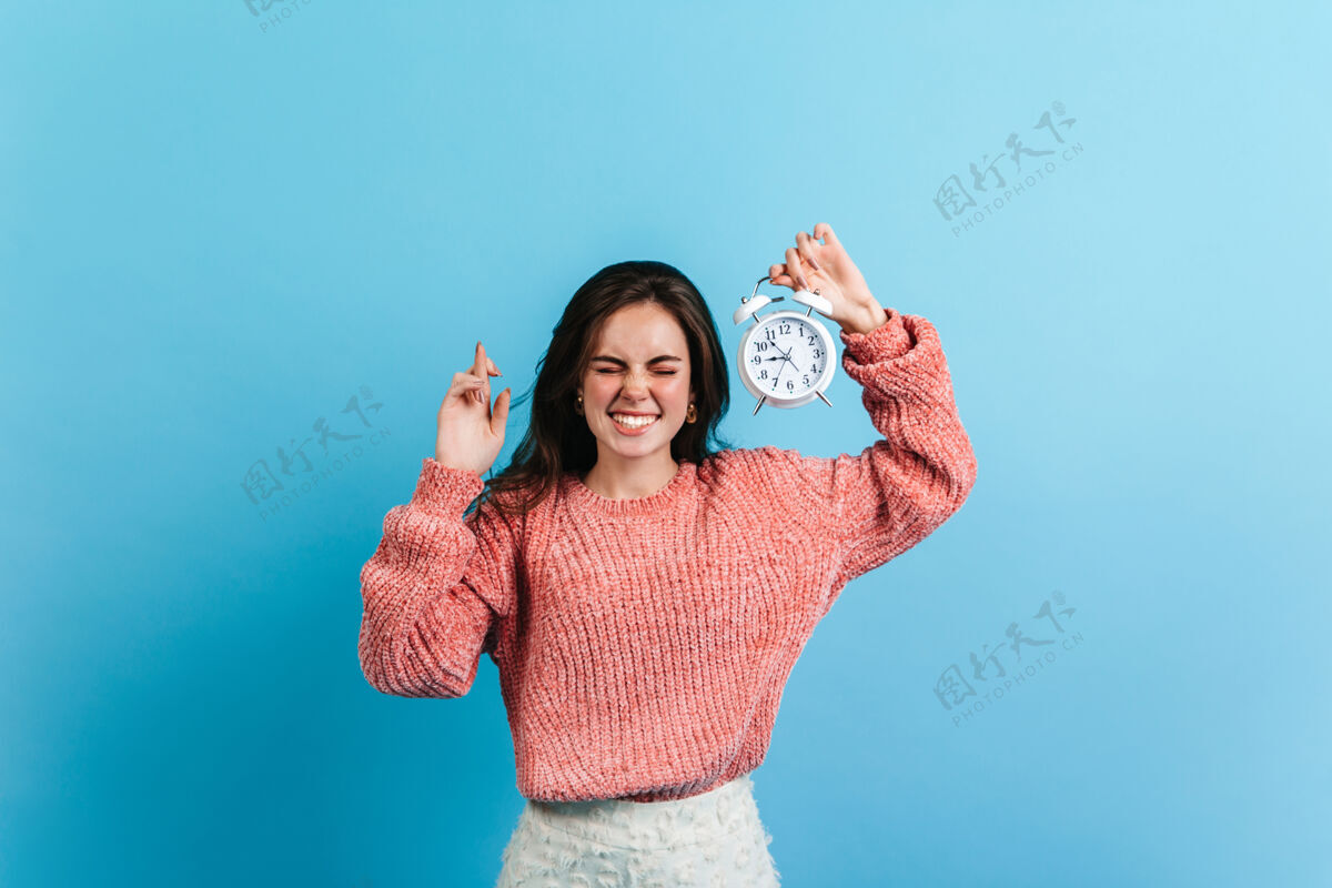 微笑深色头发的女人交叉着手指 拿着白色闹钟穿着特大号毛衣的模特在蓝色的墙上摆姿势表情粉色毛衣休闲