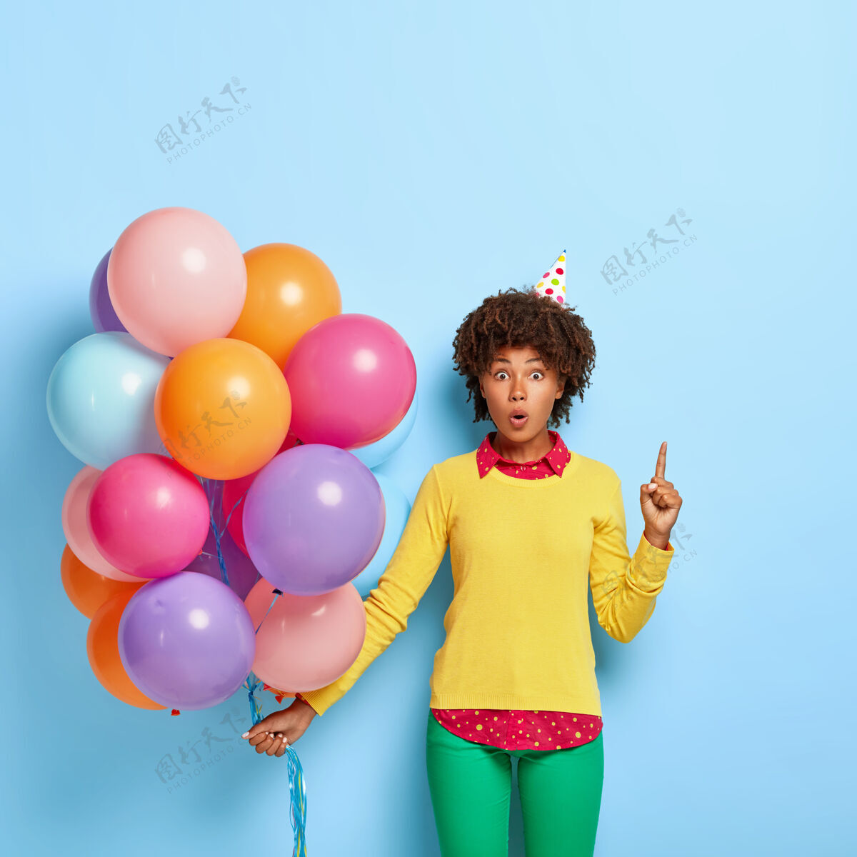 圆锥形好奇的女人拿着五颜六色的气球 穿着黄色毛衣摆姿势生日震惊人