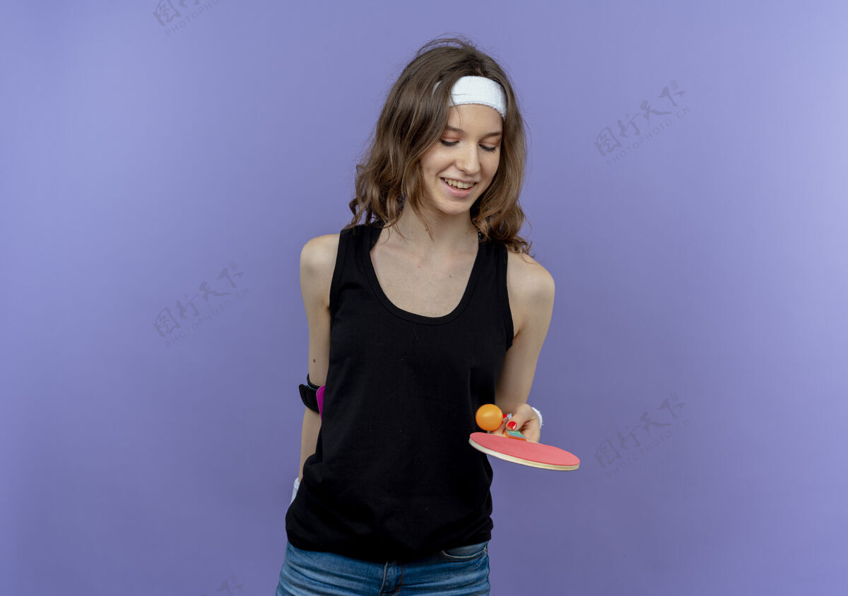 微笑身穿黑色运动服的年轻健身女孩戴着头带 手里拿着球拍和乒乓球 微笑着站在蓝色的墙上扔着球扔头带女孩