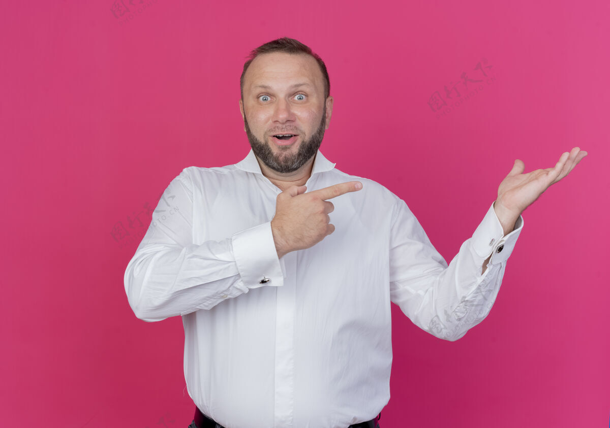 穿着一个留着胡子的男人穿着白衬衫 手臂指向一边 微笑着 站在粉色的墙上 惊讶地看着手指展示胡须