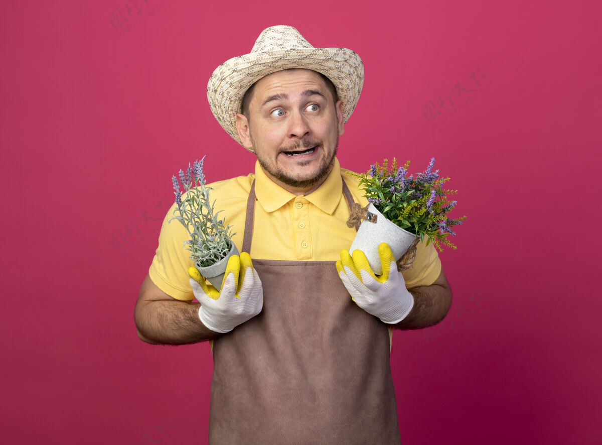 看年轻的园丁 穿着连体衣 戴着帽子 戴着工作手套 手里拿着盆栽植物 一边看一边感到困惑穿男人粉色