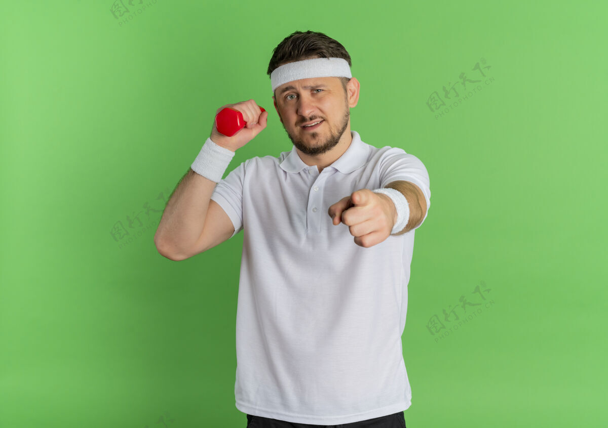 锻炼身穿白衬衫 头箍的年轻健身男子站在绿色背景上练习哑铃 手指对着摄像机哑铃男士头带