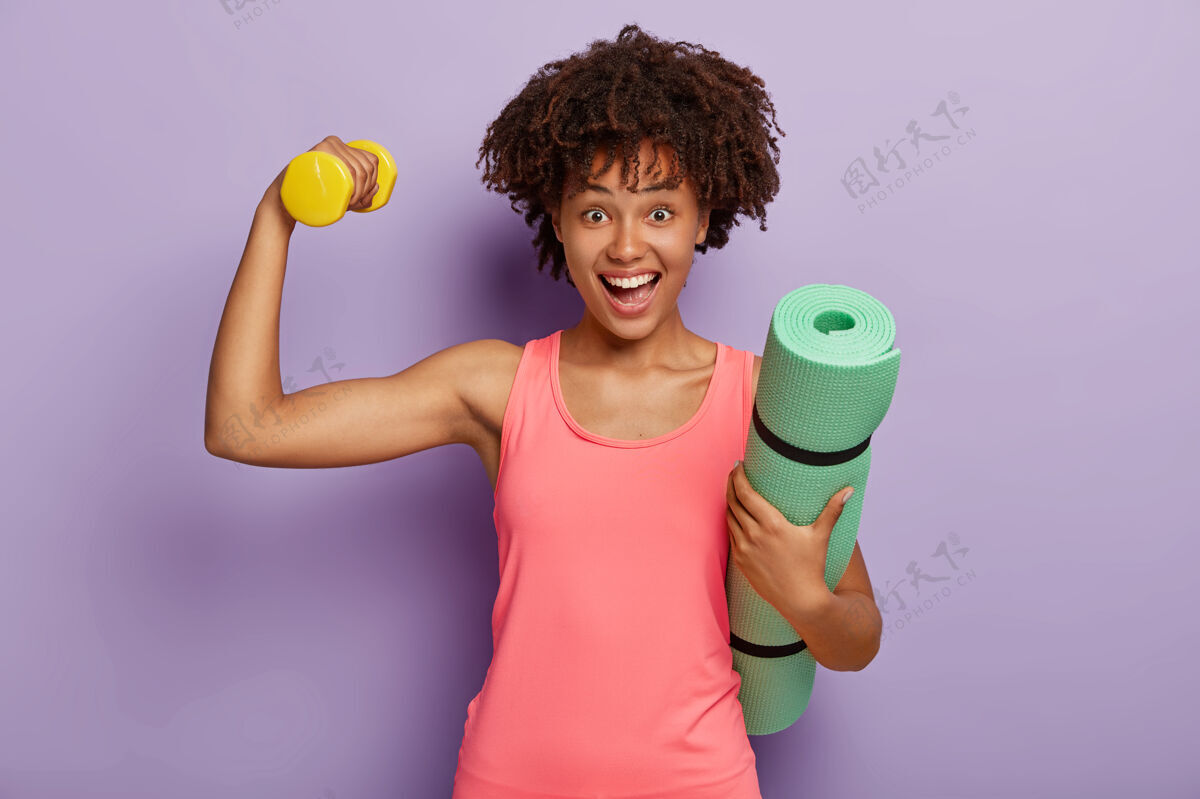 女子非洲发型快乐女人横拍 有肱二头肌举重 拎着卷起的健身垫 穿着粉色背心 神采奕奕 靠着紫色墙壁模特运动 动力形状欢呼私人教练