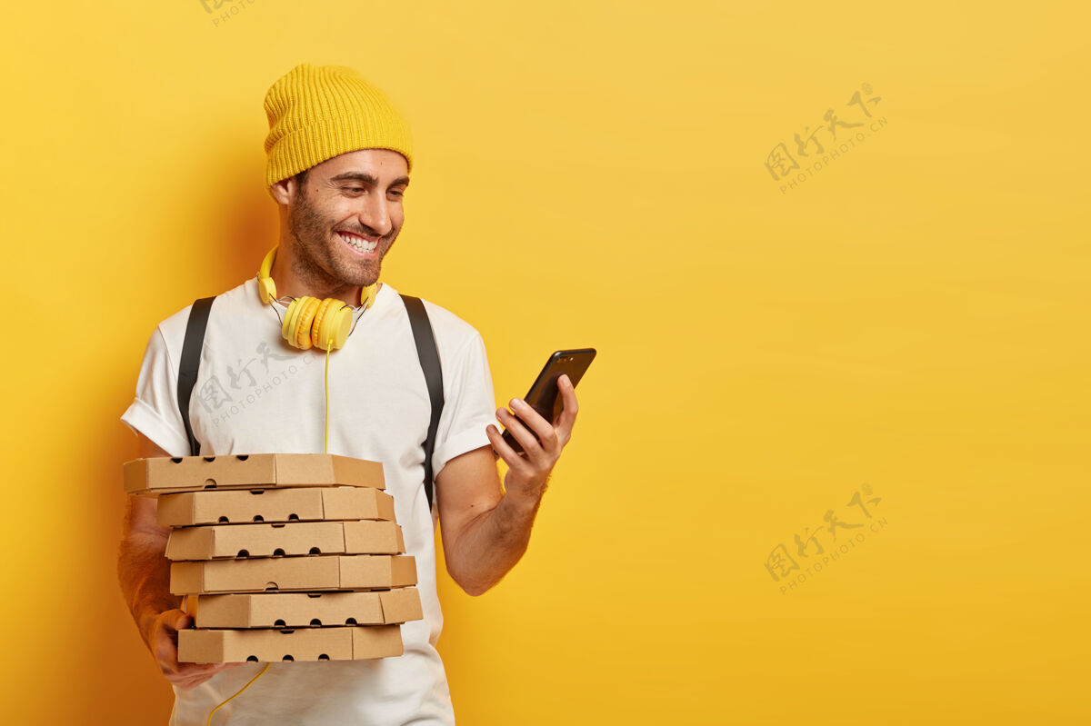 意大利人快乐男快递员的肖像通过手机查看到客户家的路线 拿着装有比萨饼的纸箱 穿着随意 通过耳机收听音频快乐电子送货