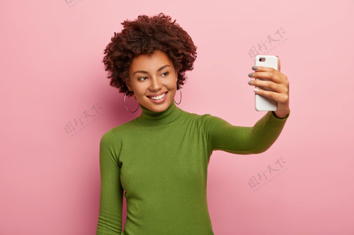 乐趣快乐的年轻女子在她的智能手机上为自己拍照 愉快地微笑着 穿着绿色高领套头衫 有着非洲式的发型 模特对着粉色的墙壁该自拍了快乐吸引力放松