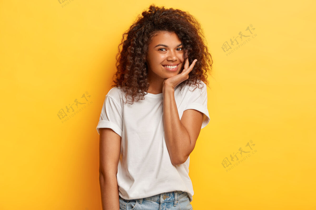 外观照片中 积极的黑皮肤女人 清爽的头发 温柔的微笑 抚摸着下巴 穿着休闲的白色t恤和牛仔裤 站在黄色的墙上人类请快乐