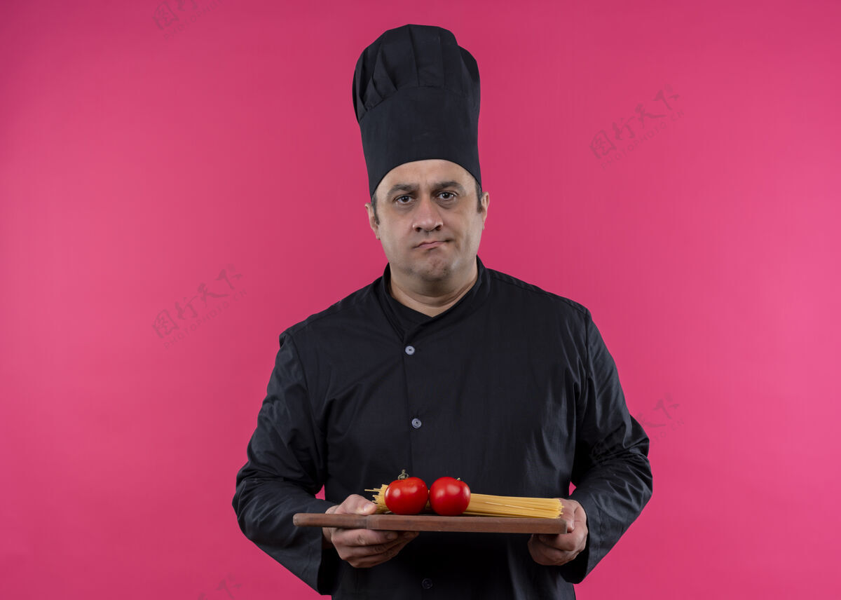 木头男厨师身穿黑色制服 头戴厨师帽 手拿西红柿木砧板 严肃的脸站在粉色背景下看着摄像机站着黑色脸