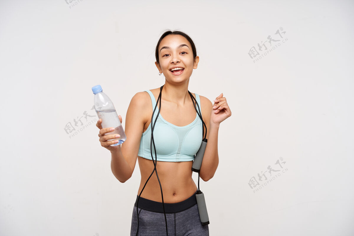 高兴快乐的棕色眼睛的年轻女子 没有化妆 一边愉快地看着前面 一边完成她的训练 一边站在白色的墙上 手里拿着一瓶水积极训练瘦身