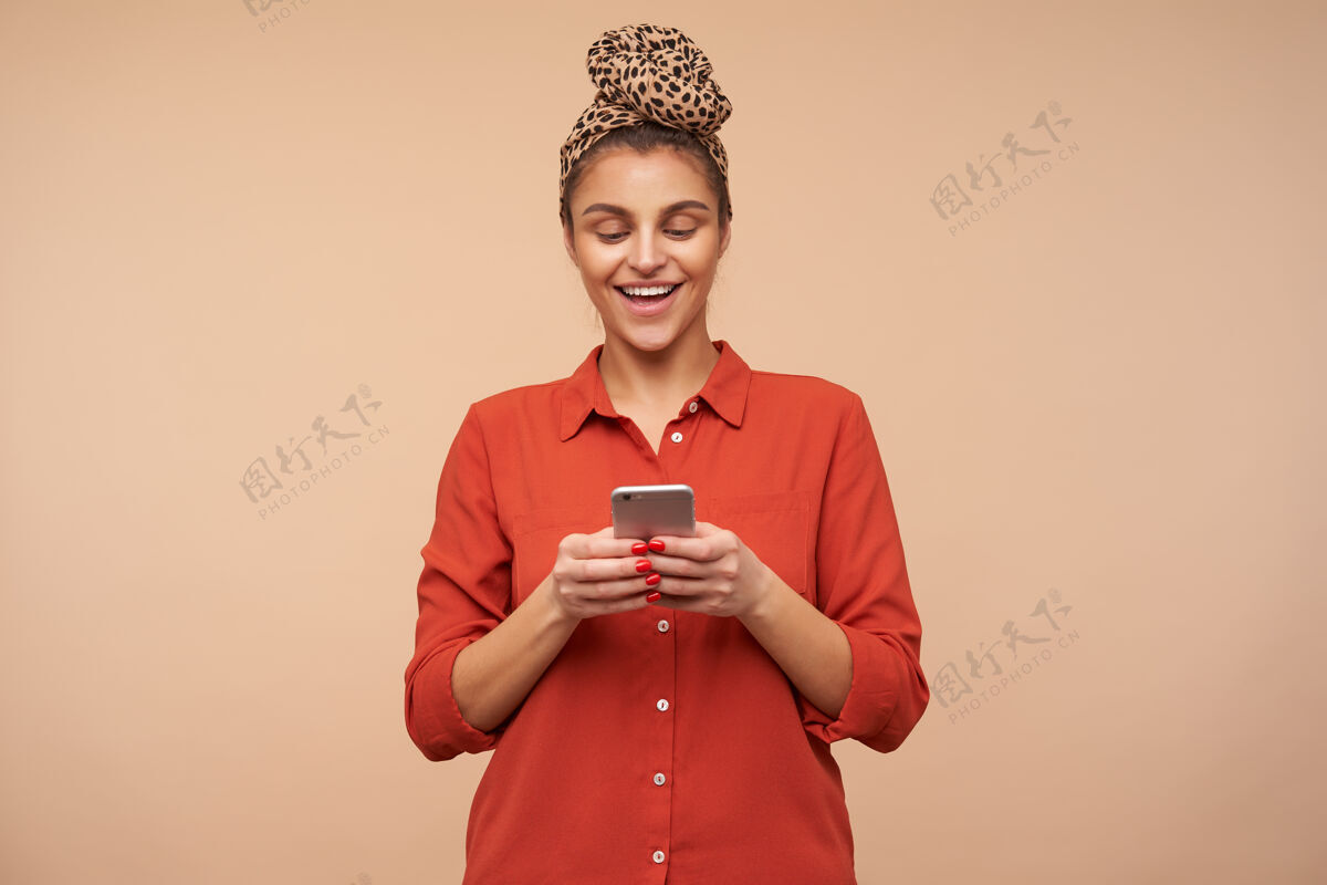 穿着快乐的年轻可爱的棕色头发的女人 戴着头巾 举着手机 高兴地看着屏幕 在米色的墙上摆姿势化妆品头巾爱