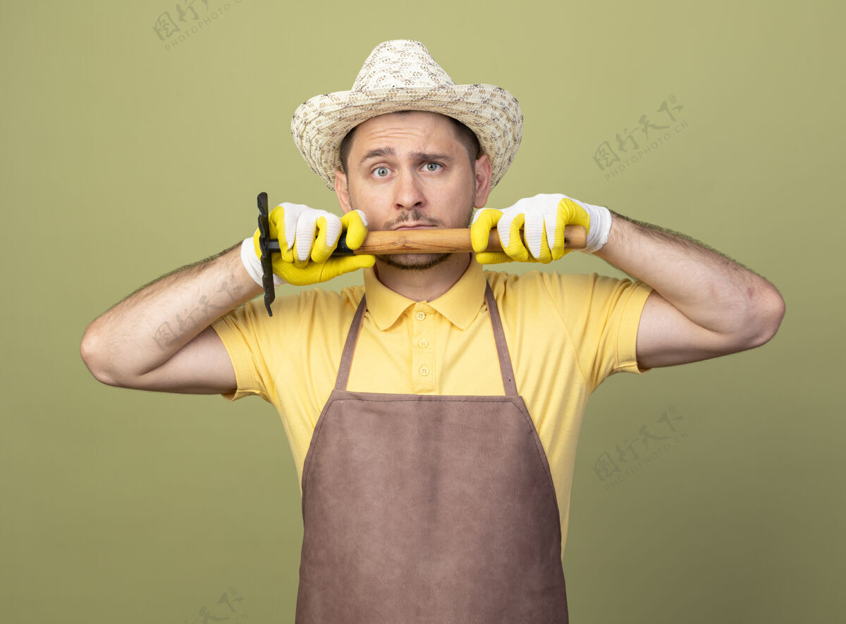 人年轻的园丁 穿着连体衣 戴着帽子 戴着工作手套 手里拿着小耙子 看着前面 严肃的脸站在明亮的墙上脸人花园