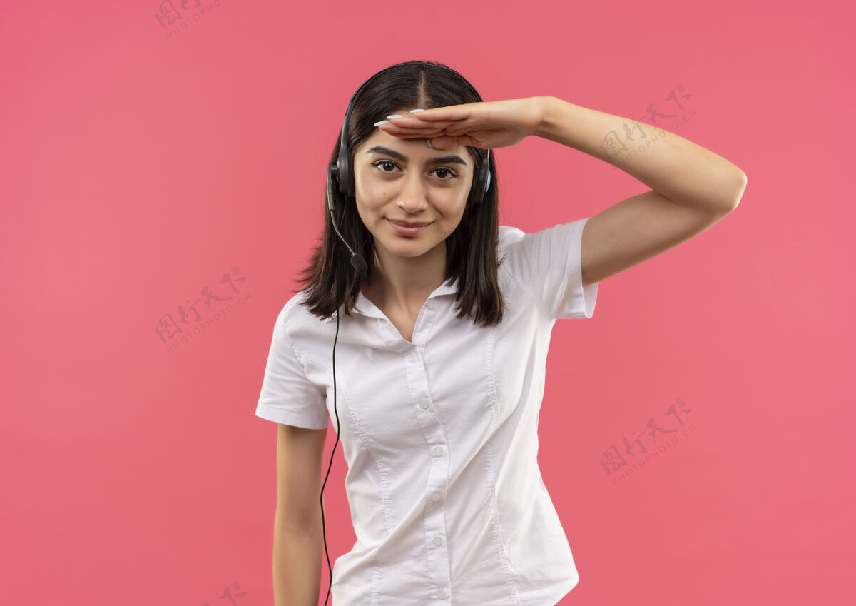 站穿着白衬衫戴着耳机的年轻女孩 手举着头站在粉红色的墙上远远望着年轻衬衫头