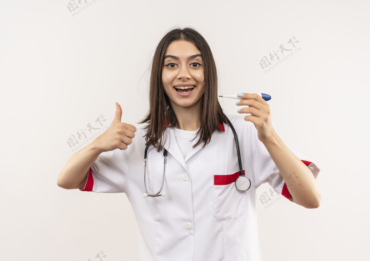 外套年轻的女医生穿着白大褂 脖子上戴着听诊器 手持数字体温计 微笑着竖起大拇指站在白墙上表演人听诊器