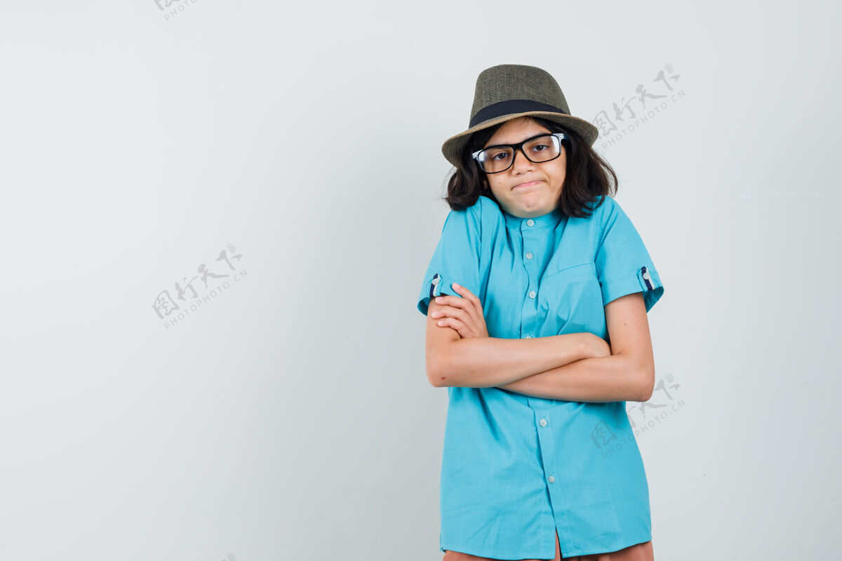 看年轻的女士交叉双臂站在蓝色衬衫 帽子 眼镜 看起来没有头脑帽子手臂室内