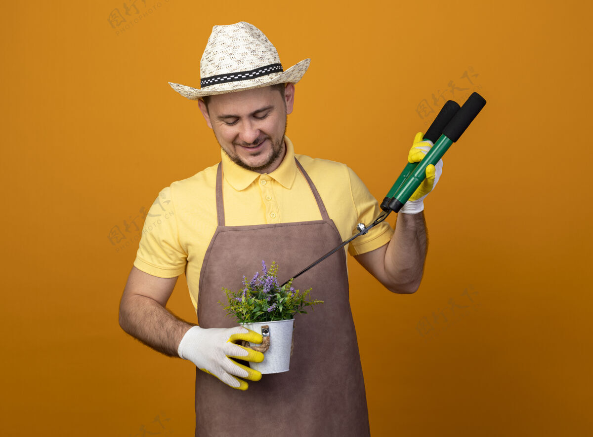 连身衣年轻的园丁穿着连体衣 戴着帽子 拿着树篱剪和盆栽植物 站在橙色的墙上微笑着看着它橙色微笑人