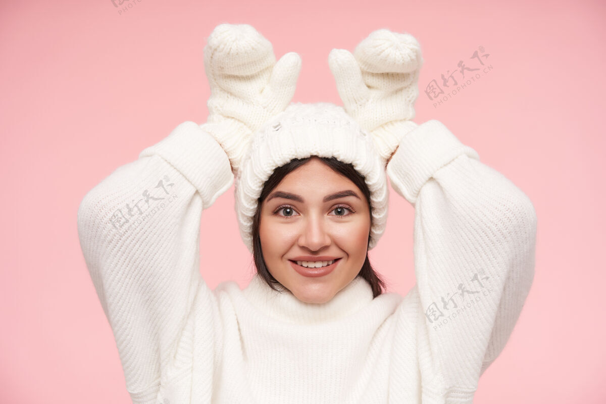 年轻快乐的年轻迷人的棕色头发的女人穿着白色毛衣 戴着手套和帽子 在粉红色的墙上摆姿势 举起手来模仿发髻的耳朵发型兔子穿