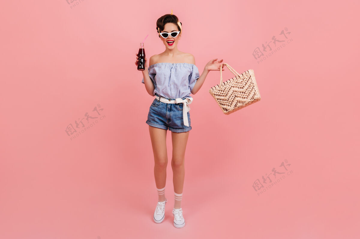 孤立的粉红色戴着太阳镜的小女孩的全貌穿着考究的年轻女子拿着苏打水和包的摄影棚照片时尚行头饮料