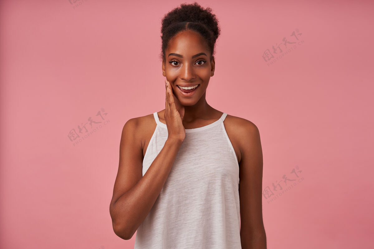 发型快乐的年轻漂亮的黑发女士 随意的发型 情绪化地举起她的手 开心地微笑着看着前面 在粉红色的墙上摆姿势女人衬衫女性