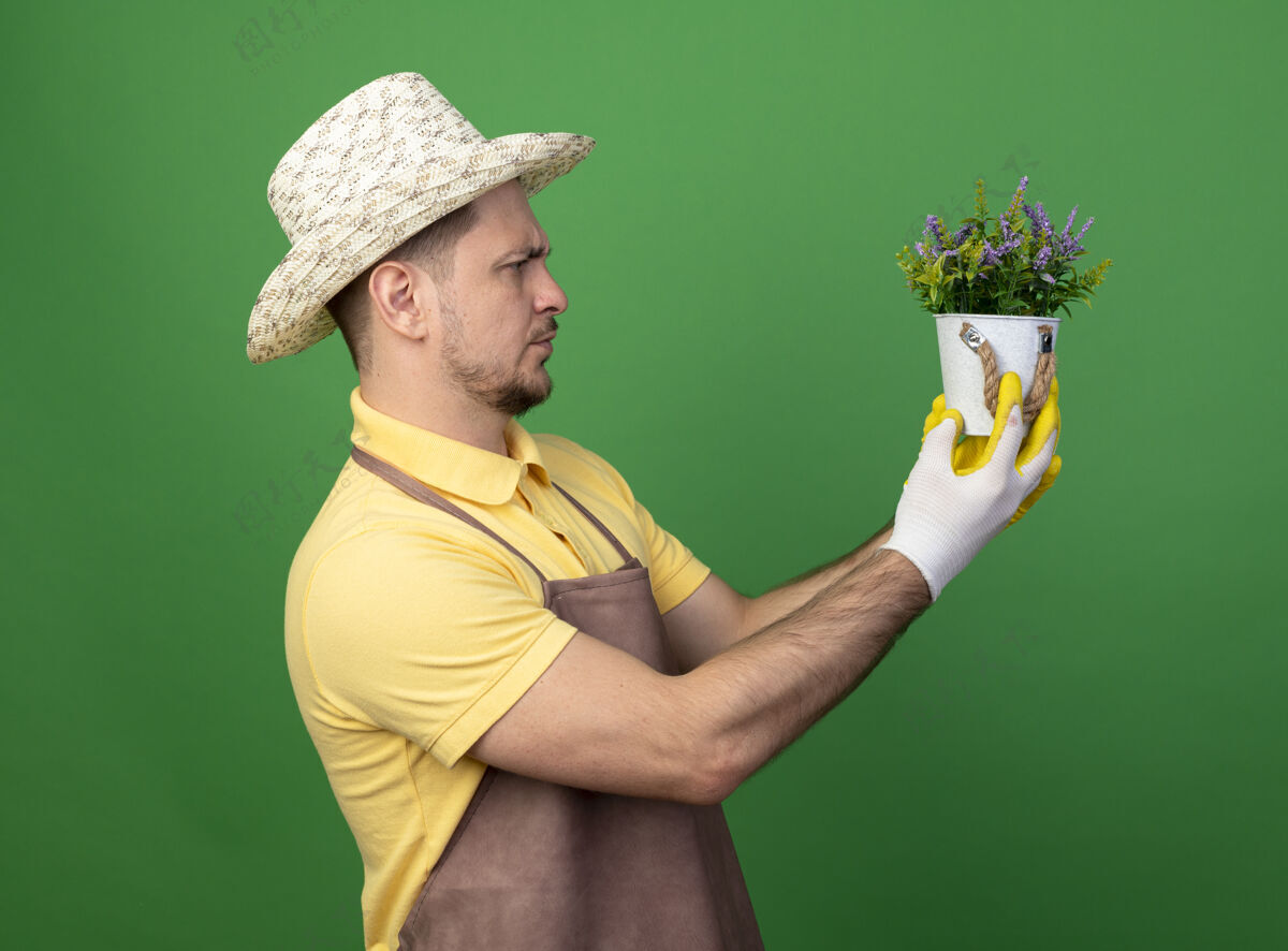 人年轻的园丁穿着连体衣 戴着帽子 戴着工作手套 手里拿着盆栽植物 站在绿色的墙上严肃地看着它绿色站着制服