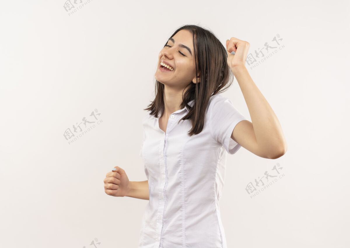 人穿着白衬衫的年轻女孩快乐而积极地闭着眼睛站在白墙上手势眼睛闭合