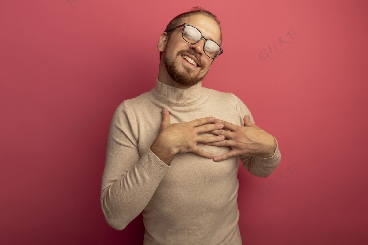 抱着身穿米色高领毛衣 戴着眼镜的年轻帅哥 站在粉色的墙上 手放在胸前 愉快地微笑着 心怀感激眼镜帅气手